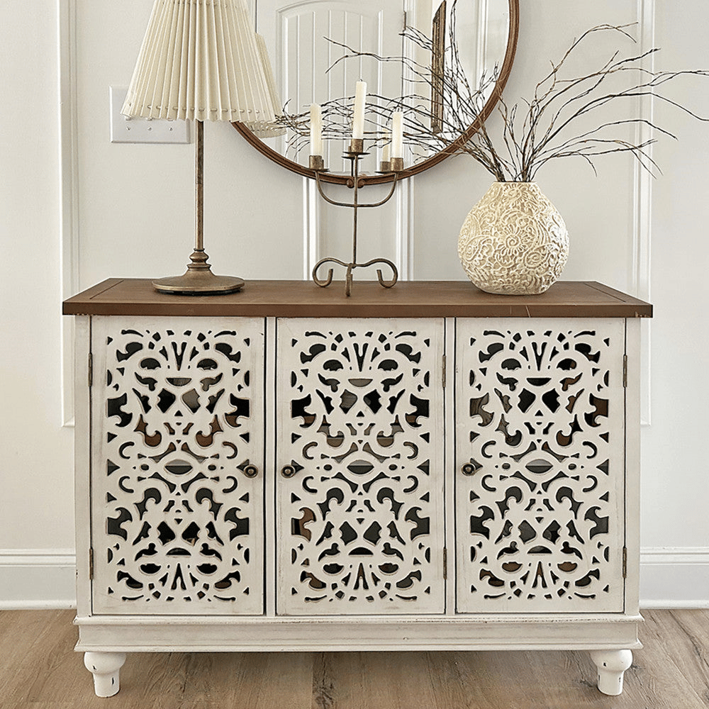 An intricately carved white sideboard with a wooden top is adorned with a pleated lampshade, a branch-filled ornate vase, and a candelabra, set against a round wall mirror.