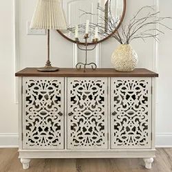 An intricately carved white sideboard with a wooden top is adorned with a pleated lampshade, a branch-filled ornate vase, and a candelabra, set against a round wall mirror.