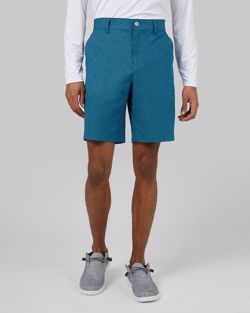 A man wearing teal shorts, a white long-sleeved shirt, and gray slip-on shoes.