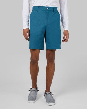 A man wearing teal shorts, a white long-sleeved shirt, and gray slip-on shoes.