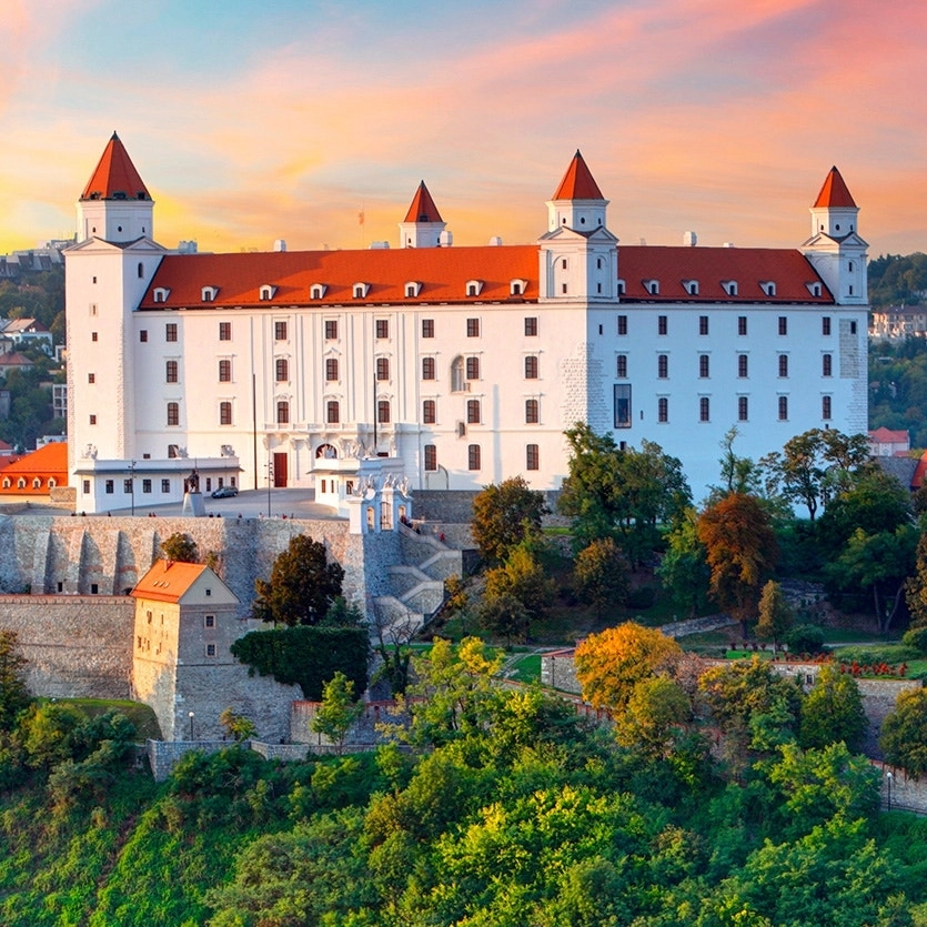 A grand white castle with red roofed towers, surrounded by greenery, against a backdrop of a sunset sky.