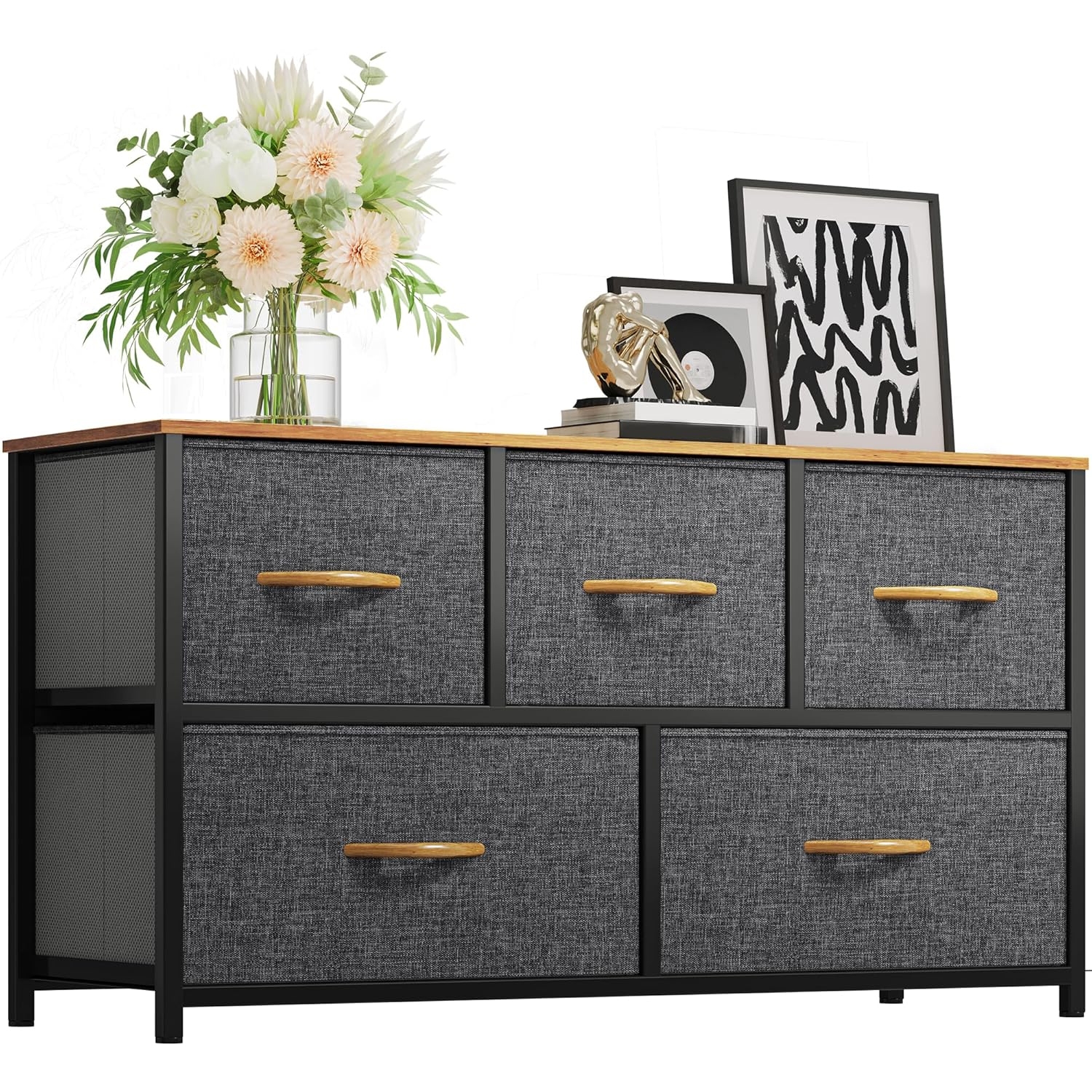A black, five-drawer fabric dresser with wooden handles and top, decorated with a vase of flowers, framed art, and a decorative item.