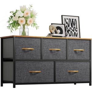 A black, five-drawer fabric dresser with wooden handles and top, decorated with a vase of flowers, framed art, and a decorative item.