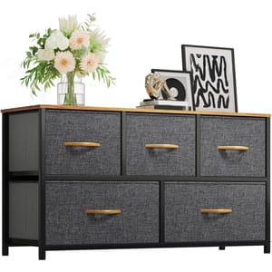 A black, five-drawer fabric dresser with wooden handles and top, decorated with a vase of flowers, framed art, and a decorative item.