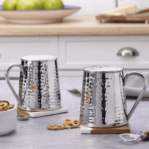 Two silver, hammered beer mugs with handles, frothy tops, and wooden coasters are on a kitchen counter beside snacks and a bottle opener.