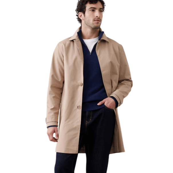A man is wearing a beige trench coat over a navy sweater and white shirt, paired with dark rolled-up jeans and brown Chelsea boots.