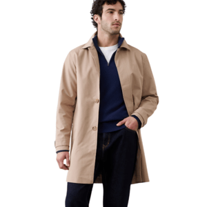 A man is wearing a beige trench coat over a navy sweater and white shirt, paired with dark rolled-up jeans and brown Chelsea boots.