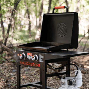 Blackstone outdoor propane griddle with a hood and two control knobs, set up in a wooded area.