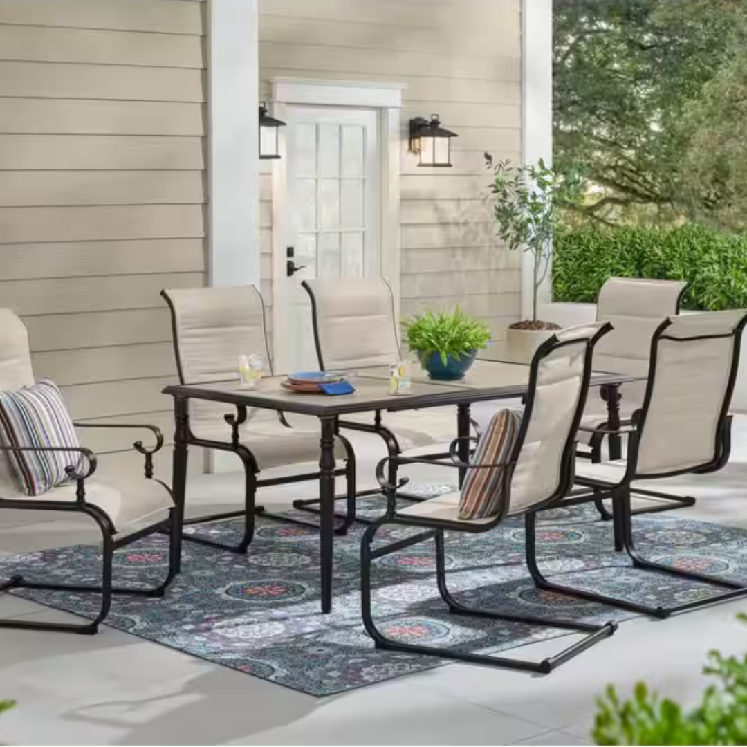 Outdoor patio set with six cushioned chairs and a rectangular table on a decorative rug. The setting is against a house with beige siding, a glass door, wall lanterns, and potted plants.