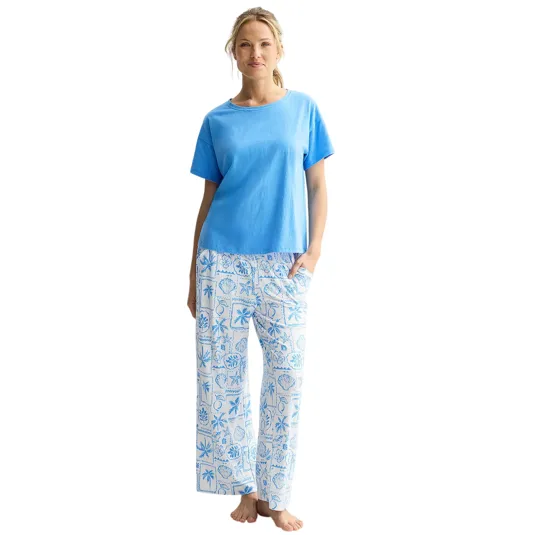 A woman is wearing a light blue short-sleeve top paired with white pajama pants featuring a light blue seashell and palm tree print.