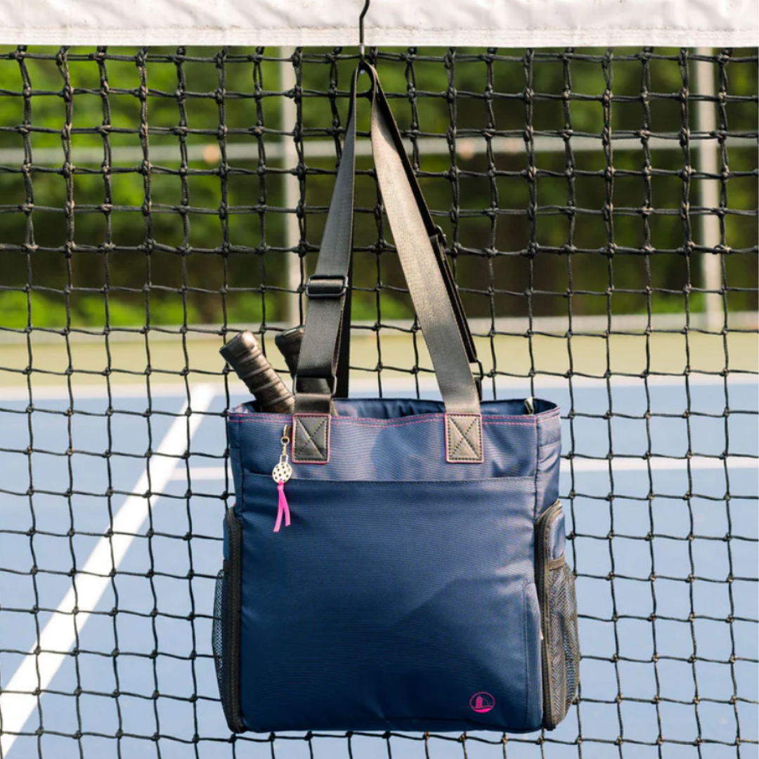 A blue tote bag with black straps is hanging on a tennis net, holding tennis rackets.