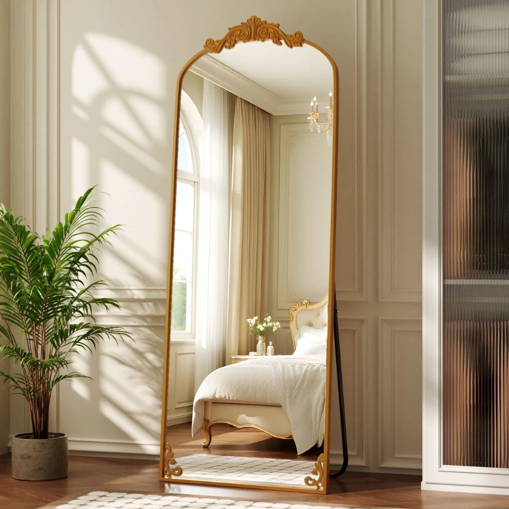 A large, ornate floor mirror with a gold frame stands in a bright bedroom, reflecting a double bed with elegant detailing. A potted plant is placed nearby on hardwood flooring, and sheer curtains cover a tall window.