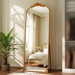 A large, ornate floor mirror with a gold frame stands in a bright bedroom, reflecting a double bed with elegant detailing. A potted plant is placed nearby on hardwood flooring, and sheer curtains cover a tall window.