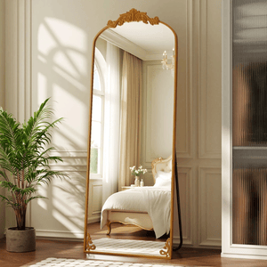 A large, ornate floor mirror with a gold frame stands in a bright bedroom, reflecting a double bed with elegant detailing. A potted plant is placed nearby on hardwood flooring, and sheer curtains cover a tall window.