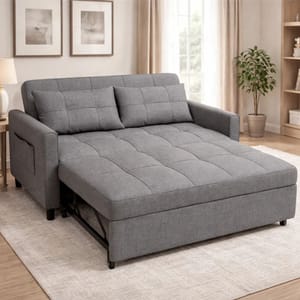 A convertible grey sofa bed with a quilted design is shown, featuring cushions and a pull-out section, placed in a living room setting with artwork, a lamp, and a potted plant.