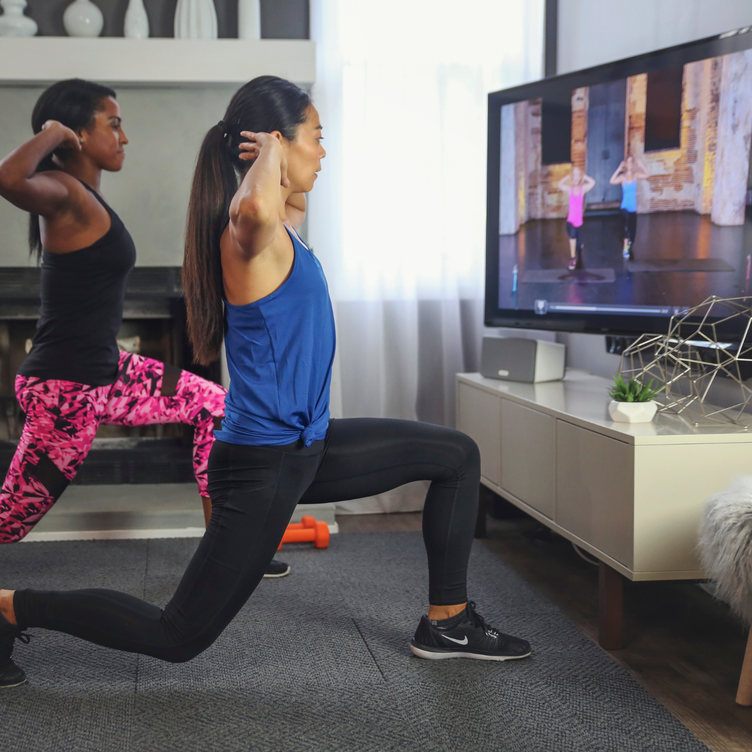 Two women perform lunges while following a workout video on TV in a living room with exercise equipment like dumbbells in the background.