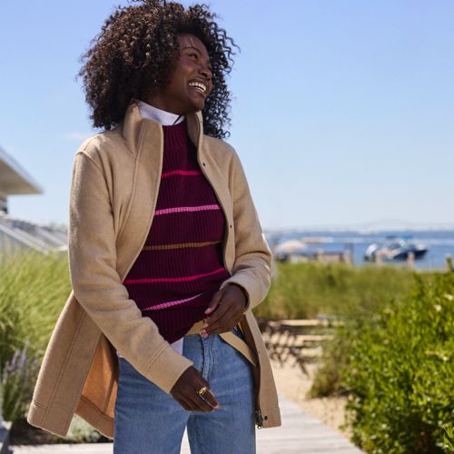 A woman is wearing a beige fleece coat with a casual cut, featuring a high collar, button closure, and side pockets. She's also wearing a striped turtleneck sweater and jeans.