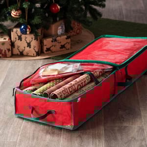 A red bag organizer with green trim for storing Christmas wrapping paper and accessories is shown under a decorated Christmas tree. It holds rolls of wrapping paper and ribbons.
