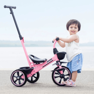 A child stands next to a pink tricycle with black wheels and a push handle.