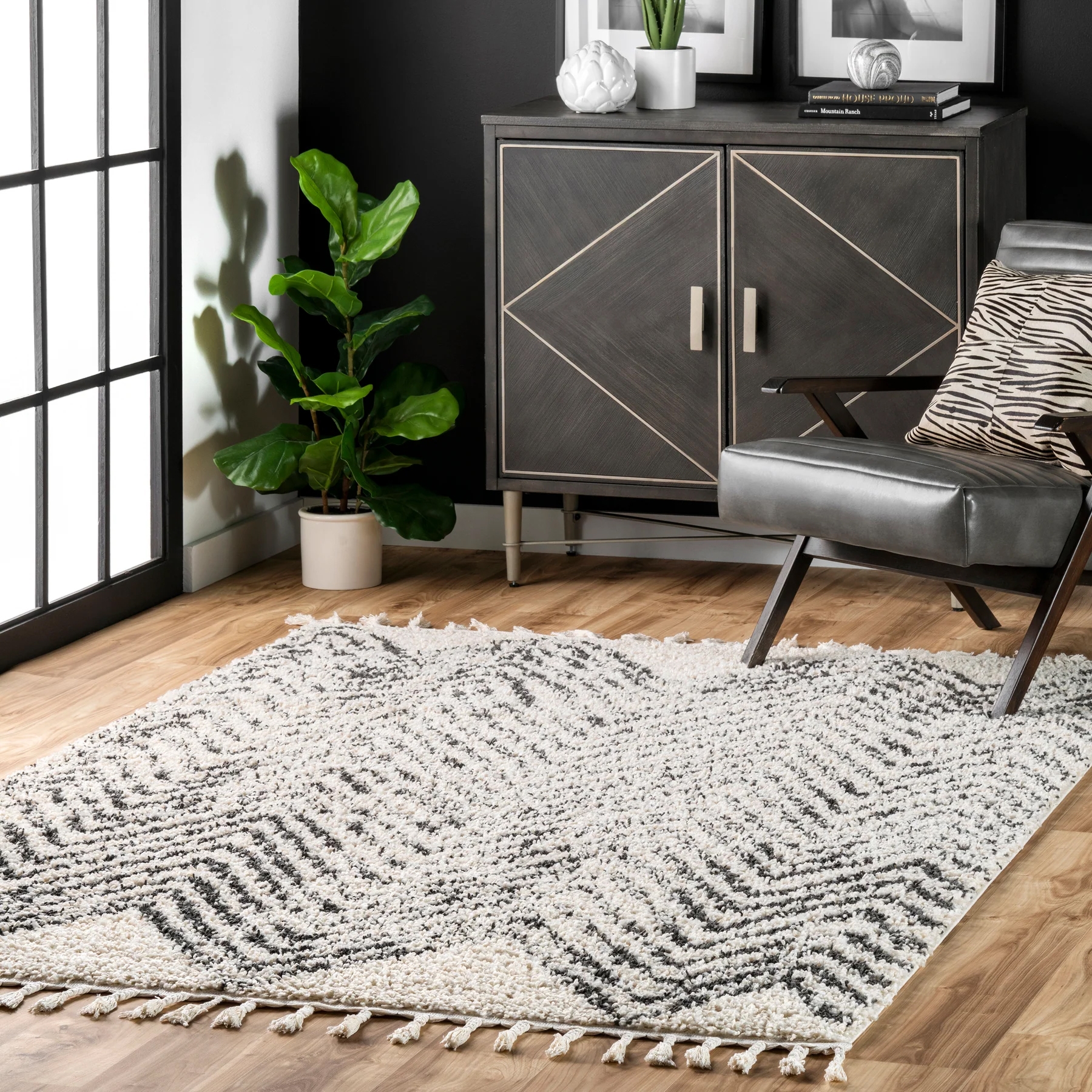 NuLoom Amanda Chevron Tassel Area Rug features a textured black and white chevron pattern with tasseled edges, set on a hardwood floor in a modern room with a sleek cabinet, a potted plant, and a gray chair with a zebra-patterned pillow.