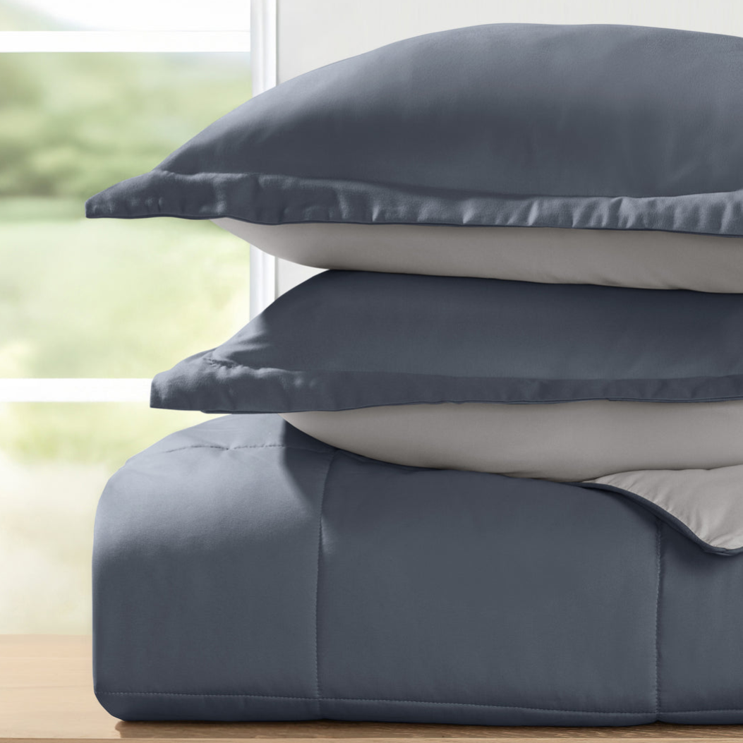 A stack of two blue and gray pillows is placed on a blue comforter, set against a blurred background seen through a window.