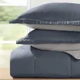 A stack of two blue and gray pillows is placed on a blue comforter, set against a blurred background seen through a window.