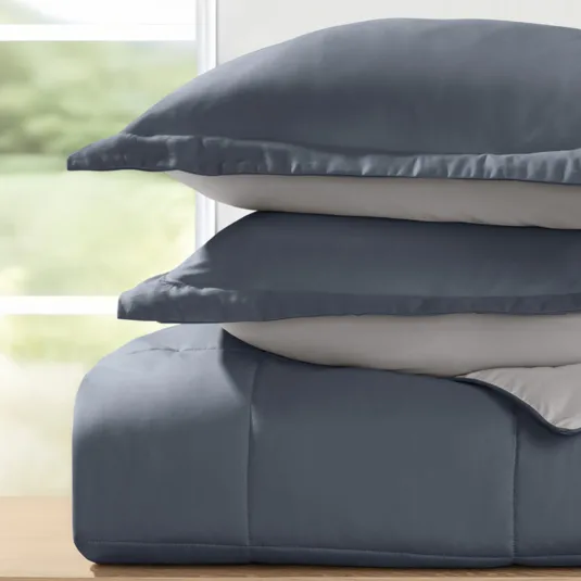 A stack of two blue and gray pillows is placed on a blue comforter, set against a blurred background seen through a window.