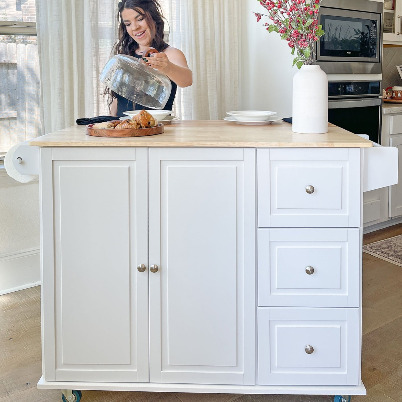 A white kitchen island with drawers and cabinets is shown, topped with a wooden surface, a large white vase with flowers, and pastries on a platter.
