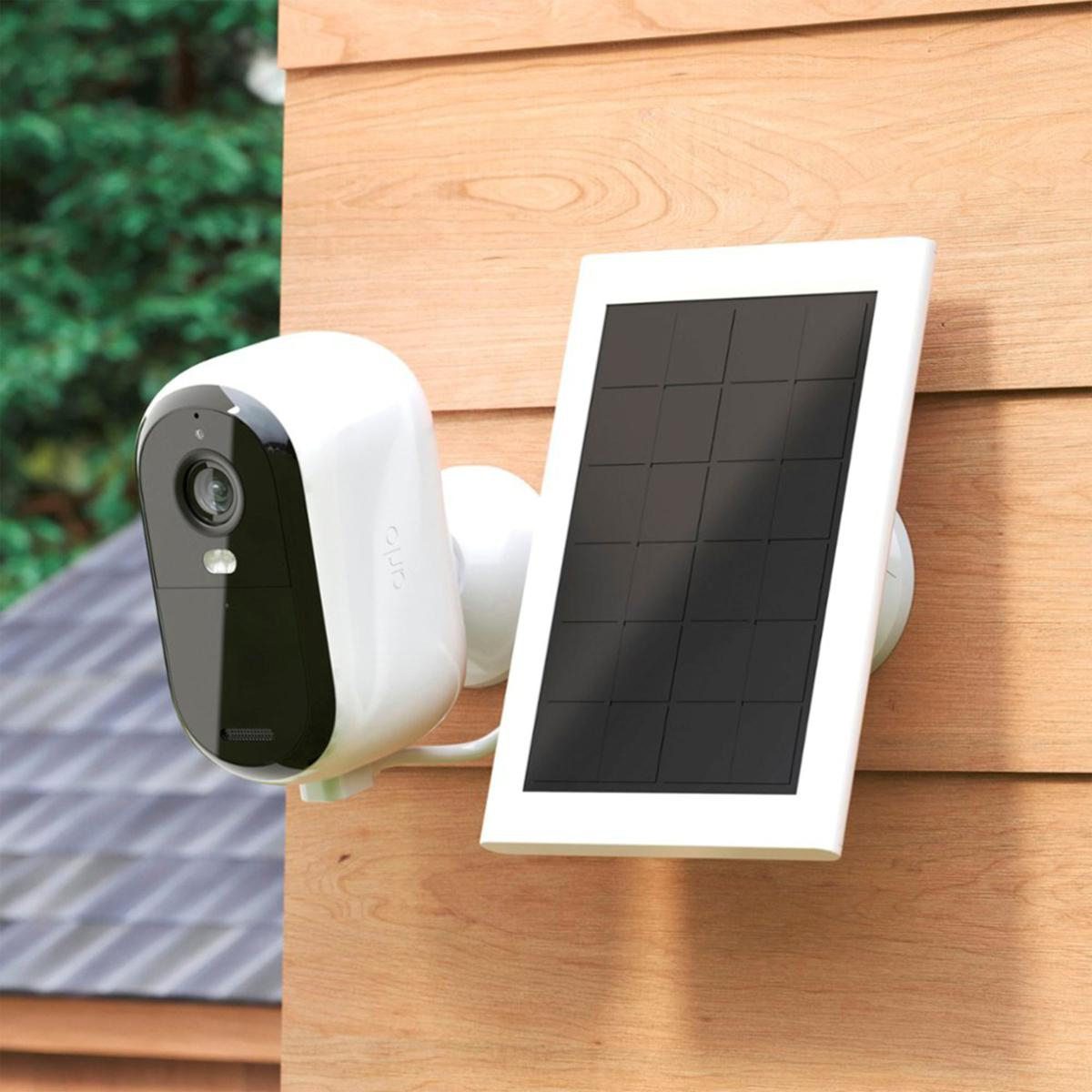 A white security camera with a black face and lens is mounted next to a rectangular solar panel on a wooden surface.