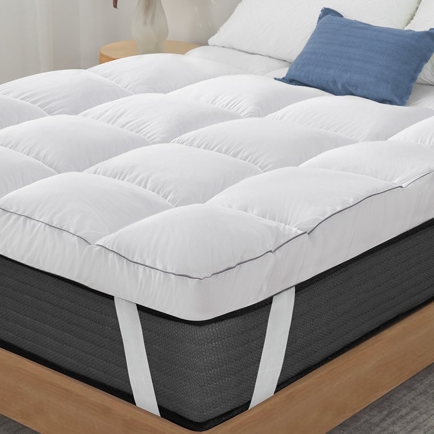 A white quilted mattress topper with corner straps is fitted over a dark-colored mattress, placed on a wooden bed frame. A blue pillow and white bedding are visible in the background.