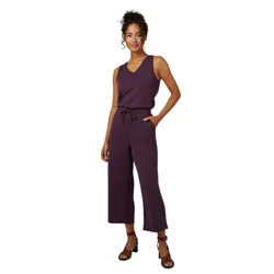 A woman is wearing a sleeveless V-neck purple jumpsuit with a drawstring waist and wide-leg cropped pants, paired with brown heeled sandals.