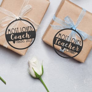 Two brown paper-wrapped gifts with decorative ribbons and circular tags reading \“Most Loved Coach\“ and \“Most Loved Teacher Johnny,\“ alongside a white rose.