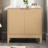 A compact, light wood storage cabinet with two vertically reeded doors, sits on short legs. It features minimalist design, a sleek top surface with books, a plant, and a vase, blending functionality with modern style.