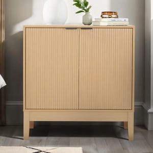 A compact, light wood storage cabinet with two vertically reeded doors, sits on short legs. It features minimalist design, a sleek top surface with books, a plant, and a vase, blending functionality with modern style.