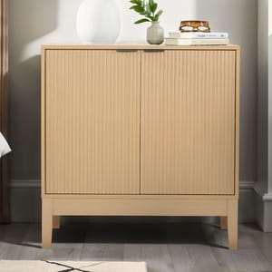 A compact, light wood storage cabinet with two vertically reeded doors, sits on short legs. It features minimalist design, a sleek top surface with books, a plant, and a vase, blending functionality with modern style.