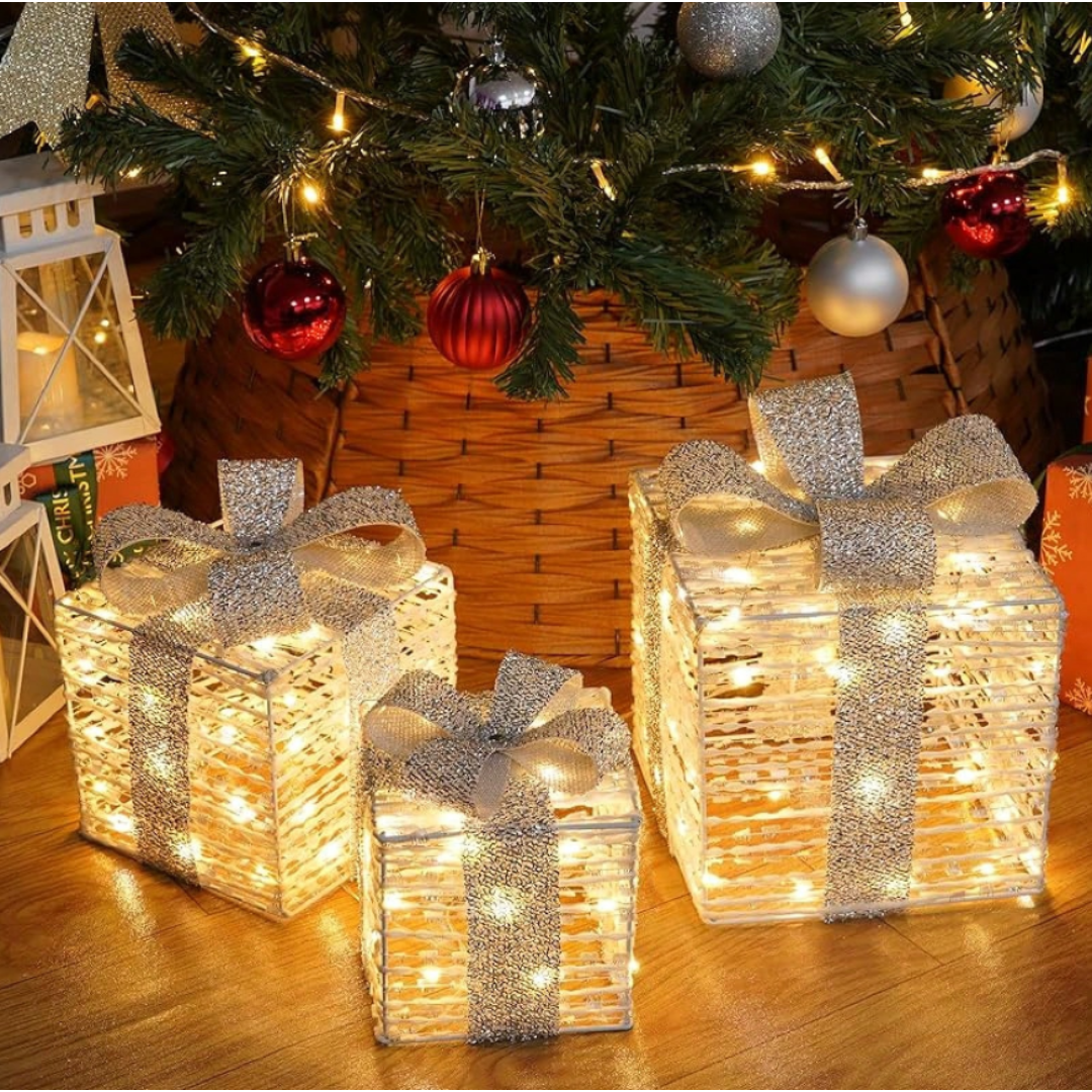 Three illuminated decorative gift boxes with silver ribbons are placed under a Christmas tree, surrounded by ornaments and holiday lanterns.