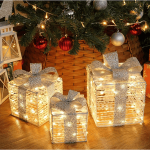 Three illuminated decorative gift boxes with silver ribbons are placed under a Christmas tree, surrounded by ornaments and holiday lanterns.