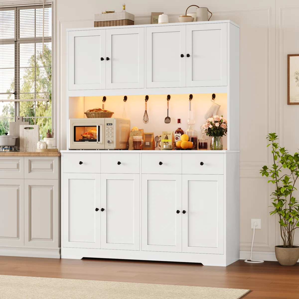 White kitchen cabinet with upper and lower storage, featuring black handles. Middle section displays a microwave and various kitchen items, including utensils and food products. Decor includes a plant, flowers, and bottles.