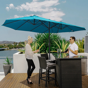 Two people stand on a wooden deck under a large blue patio umbrella. One is holding drinks near a wicker bar set, with lush plants and a scenic water view in the background.