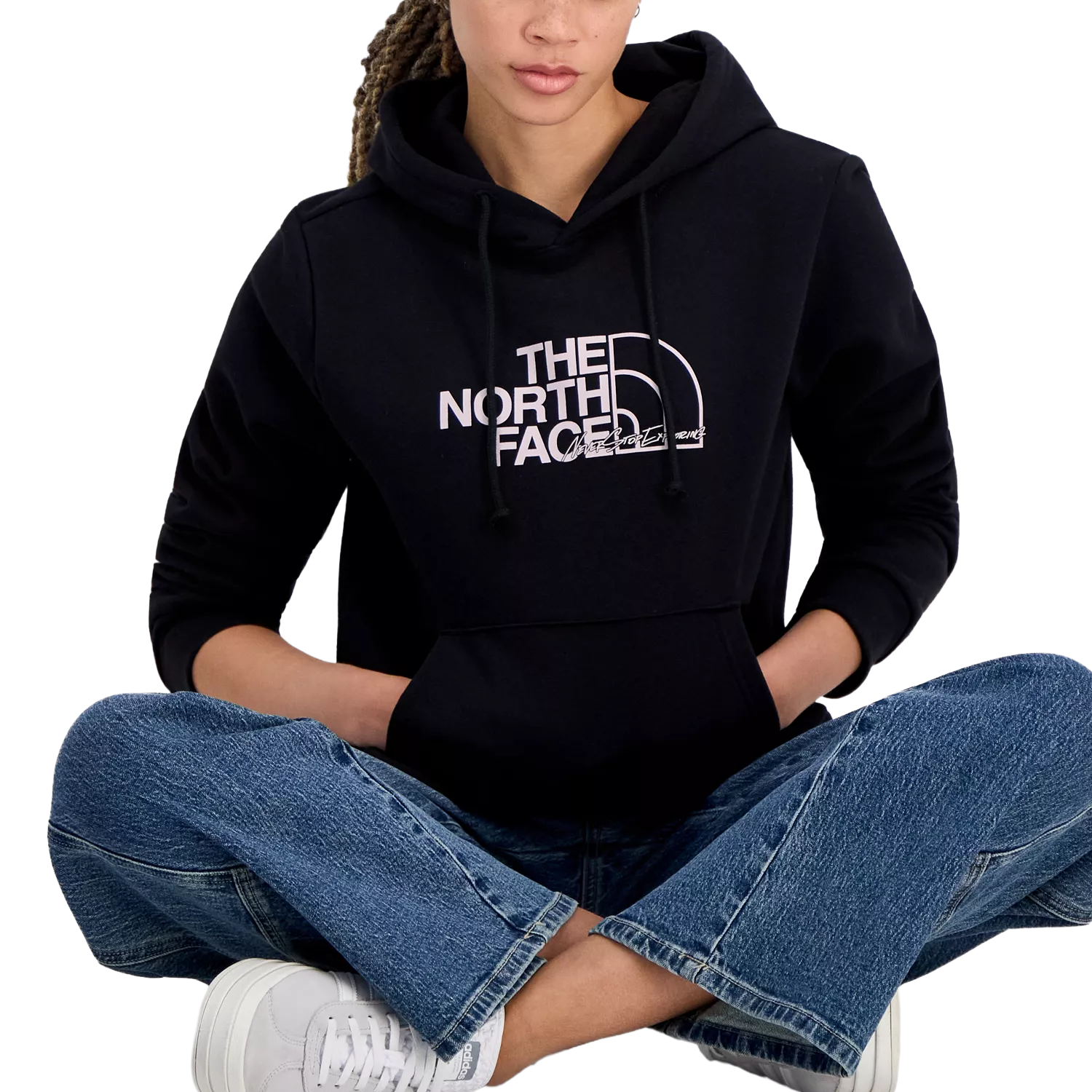 A person wearing a black The North Face hoodie with a logo on the front, paired with blue jeans and white sneakers, sitting cross-legged.