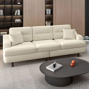 Modern living room with a beige three-seater sofa and two cushions, featuring a dark oval coffee table with decor items. Background includes a wooden bookshelf with books and ambient lighting.