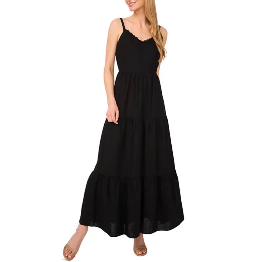 A woman is wearing a black, tiered maxi dress with thin straps and a scalloped neckline, paired with open-toed sandals.
