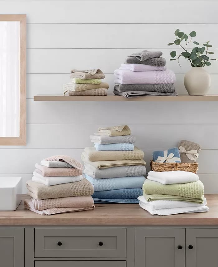 Neatly arranged, colorful stacks of towels on a wooden countertop and shelf in a bathroom setting, accompanied by a decorative vase with greenery.