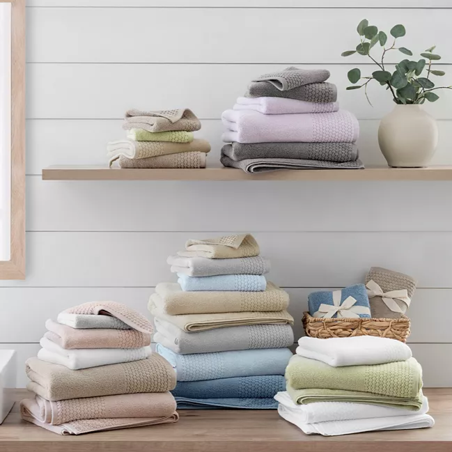 Neatly arranged, colorful stacks of towels on a wooden countertop and shelf in a bathroom setting, accompanied by a decorative vase with greenery.