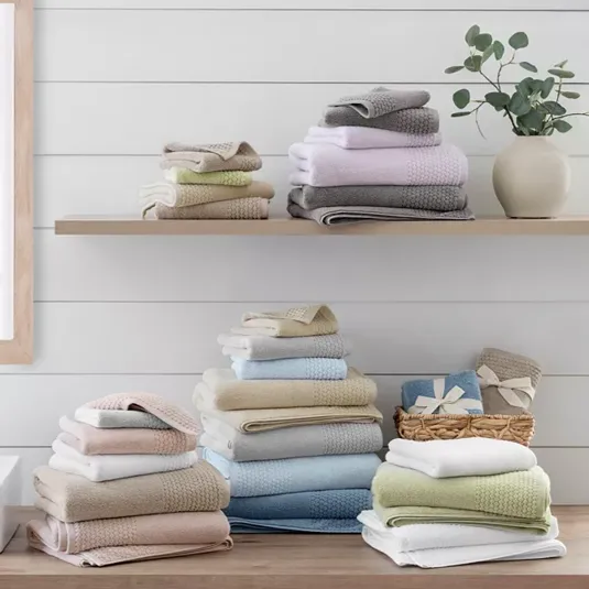 Neatly arranged, colorful stacks of towels on a wooden countertop and shelf in a bathroom setting, accompanied by a decorative vase with greenery.
