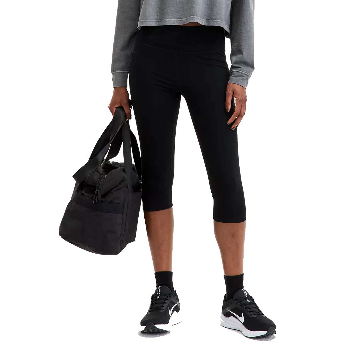 A person is wearing black cropped leggings and sneakers, holding a black duffle bag.