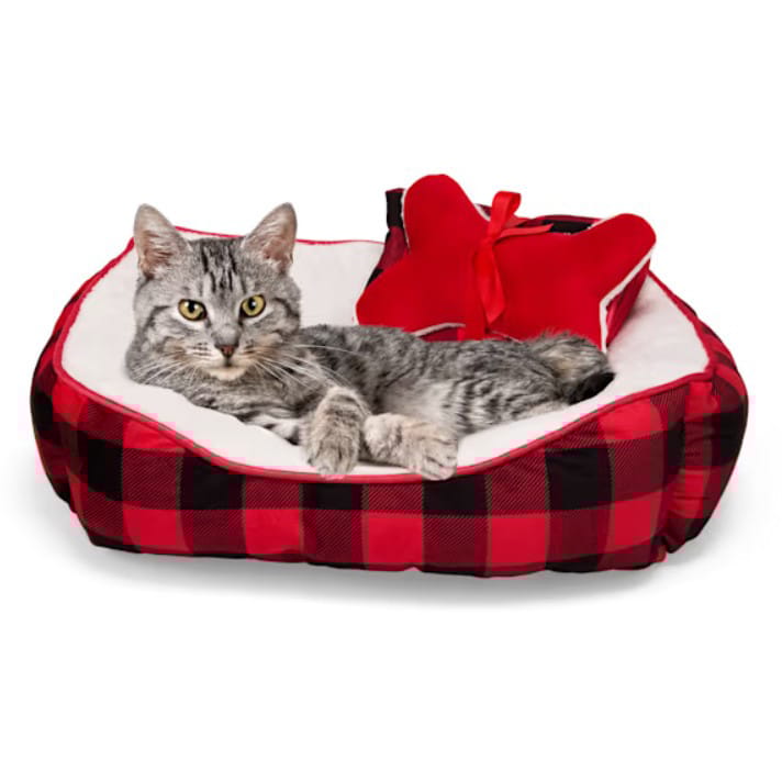 A gray tabby cat is lounging in a red and black plaid pet bed with a plush interior, accompanied by a red bow-shaped pillow.