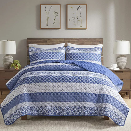 A bed with a blue and white patterned quilt and matching pillows, flanked by two bedside tables with lamps and decorative frames on the wall.
