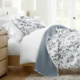 A neatly made bed with floral-patterned bedding, featuring a reversible quilt in blue and white with matching pillowcases. The room has a wooden nightstand, a table lamp, and a bright window in the background.