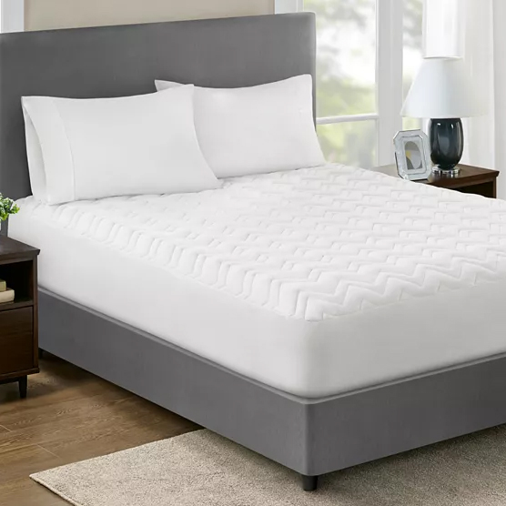 Home Design Classic Mattress Pads feature a quilted pattern, providing an extra layer of comfort and protection for mattresses. They have a sleek, white design and are shown on a bed with a grey upholstered headboard.